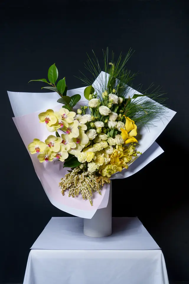 A vibrant, professionally arranged bouquet featuring yellow orchids, white roses, yellow ranunculus, and a yellow calla lily. The bouquet is wrapped in white and light pink paper, accented with green foliage and pine needles. It stands in a white cylindrical vase on a white tablecloth against a dark, plain background.