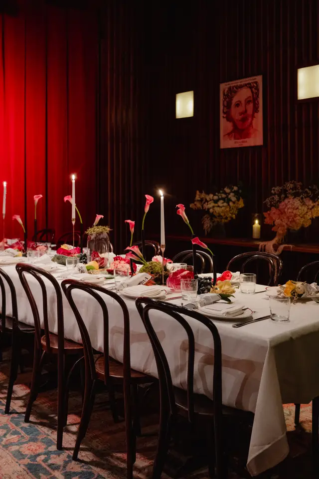 A long dining table is elegantly set for an event in a dimly lit room. The table features a white tablecloth, multiple place settings, lit candles, and various floral and fruit arrangements. The background includes rich red curtains on the left and dark wood paneling with wall sconces and a framed artwork on the right.