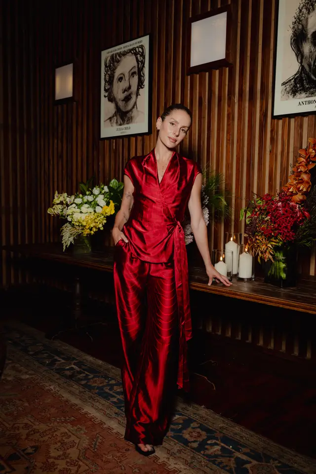 A woman in a striking red patterned satin wrap top and matching wide-leg pants stands beside a long wooden table. The table is adorned with elaborate floral arrangements featuring yellow, white, red, and orange flowers, alongside several lit pillar candles in glass holders. The background features dark wood paneling, framed portraits, and a patterned rug, creating an elegant and warm indoor event atmosphere.