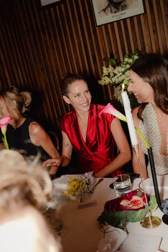 Three women are seated at a candlelit dining table adorned with floral arrangements and fruit, engaged in conversation at what appears to be an elegant event. The central woman in a red dress is smiling and looking towards another guest. The setting features warm lighting and a wooden slatted wall.