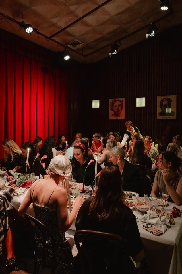 A dimly lit, warm image captures a formal dinner event with multiple guests seated around a long table. The table is elegantly set with food, drinks, white pillar candles, and pink calla lilies, creating an intimate and sophisticated atmosphere. Red curtains and dark wood paneling frame the room, with spotlights illuminating the scene.