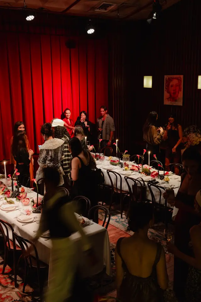 An indoor evening event is captured, showing multiple guests mingling around long, elegantly set tables. The scene is dimly lit with warm light from candles and spotlights, creating an intimate atmosphere. Rich red curtains serve as a backdrop on one side, while tables are adorned with floral arrangements, fruit, and candles.