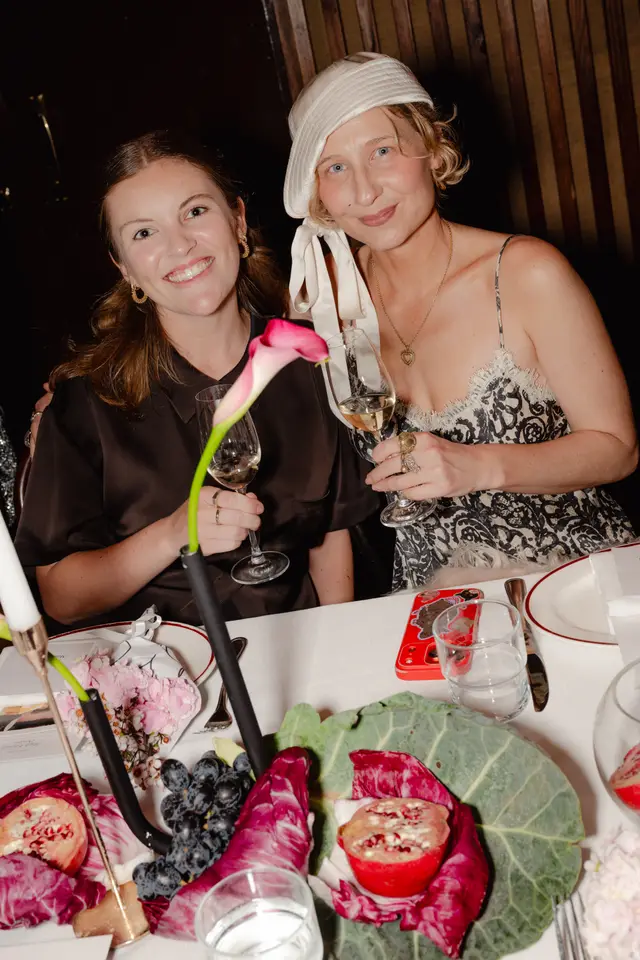 Two women are seated at a dinner table, smiling and holding wine glasses, at what appears to be a formal event. The table is elaborately decorated with fresh produce like pomegranates, grapes, and radicchio, alongside a pink calla lily and candles. The background is dark, suggesting an evening setting.