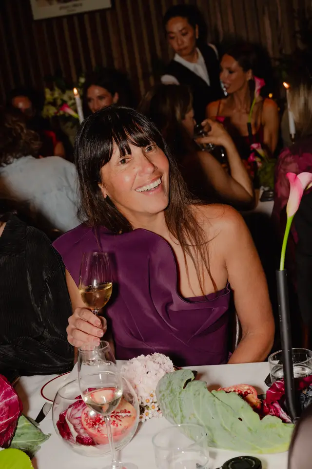A smiling woman with dark hair and a purple one-shoulder dress holds a wine glass, looking towards the camera. She is seated at a table adorned with floral arrangements, pomegranates, and radicchio, suggesting a festive or launch event. Other guests and a server are visible in the softly lit background.