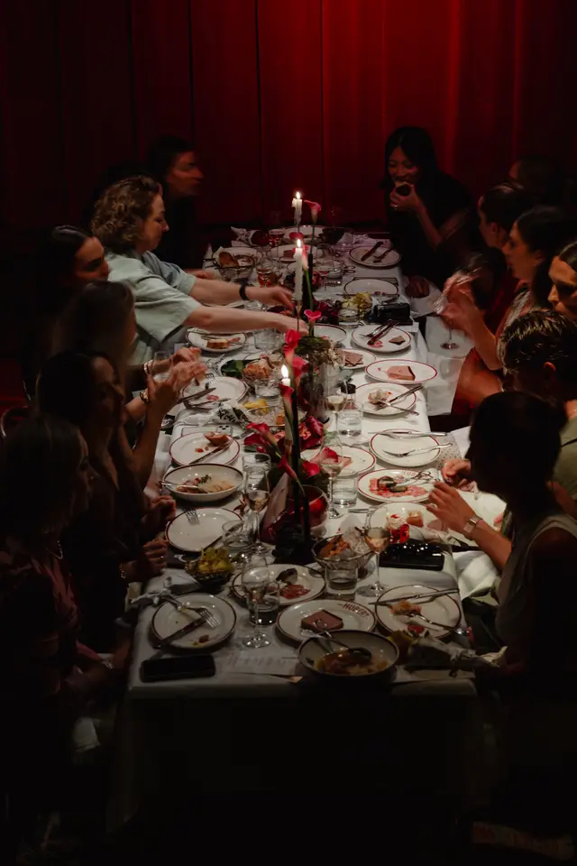 This image captures a lively, intimate dining event from an overhead perspective. Multiple guests are seated around a long table adorned with white tablecloths, flickering candles, and elegant floral arrangements featuring red calla lilies. Various dishes, wine glasses, and cutlery are visible, suggesting a multi-course meal in progress against a backdrop of dark red curtains.
