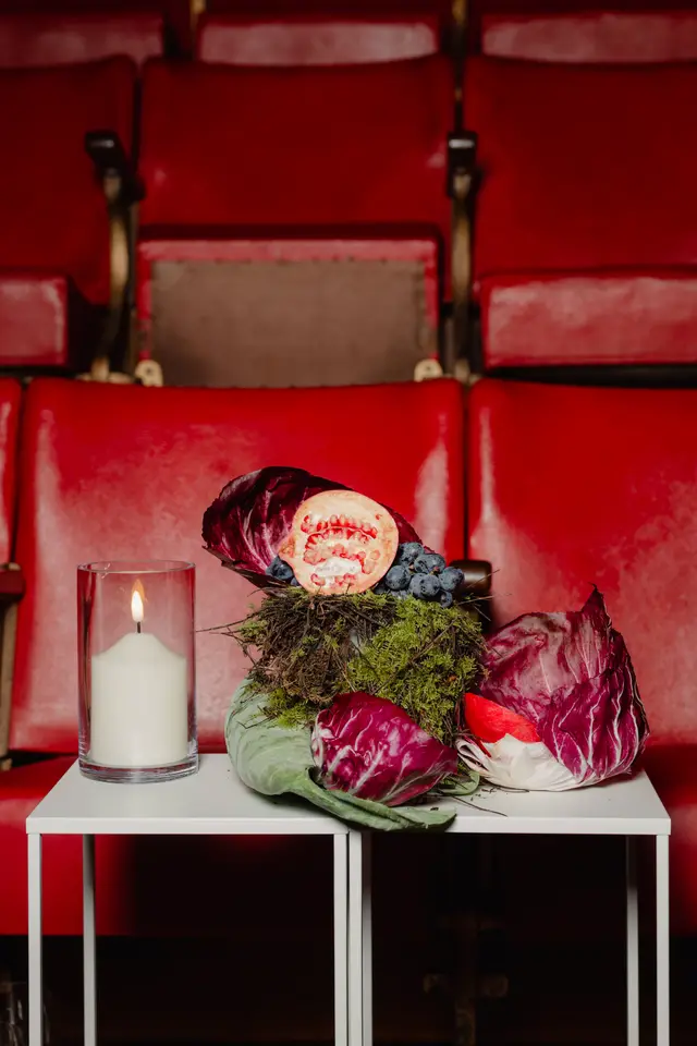 A styled still life arrangement featuring a half pomegranate, dark grapes, and various types of radicchio and cabbage leaves. The arrangement rests on a bed of moss and twigs, placed on a white table next to a lit white pillar candle in a glass holder. The background is blurred, showing rows of red upholstered theater seats.