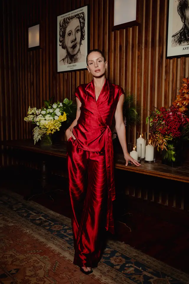 A woman in a vibrant red satin wrap top and matching wide-leg pants stands confidently, looking directly at the camera. She is positioned between two elaborate floral arrangements and lit candles on a dark wooden table. The background features a slatted wooden wall adorned with framed black and white portraits, creating a sophisticated and warm event atmosphere.