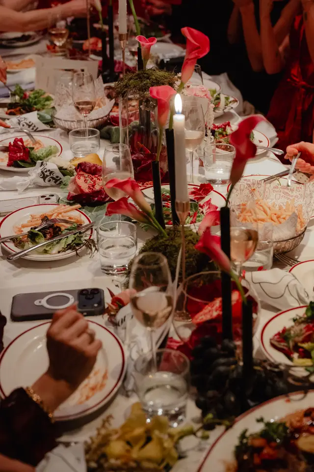A close-up, high-angle shot captures a beautifully set dinner table during an event, adorned with vibrant pink calla lilies, moss, and lit candles. Various dishes, including salads, fries, and grapes, are visible on white plates with red rims. Several hands are present, suggesting guests are actively dining in a warm, intimate atmosphere.