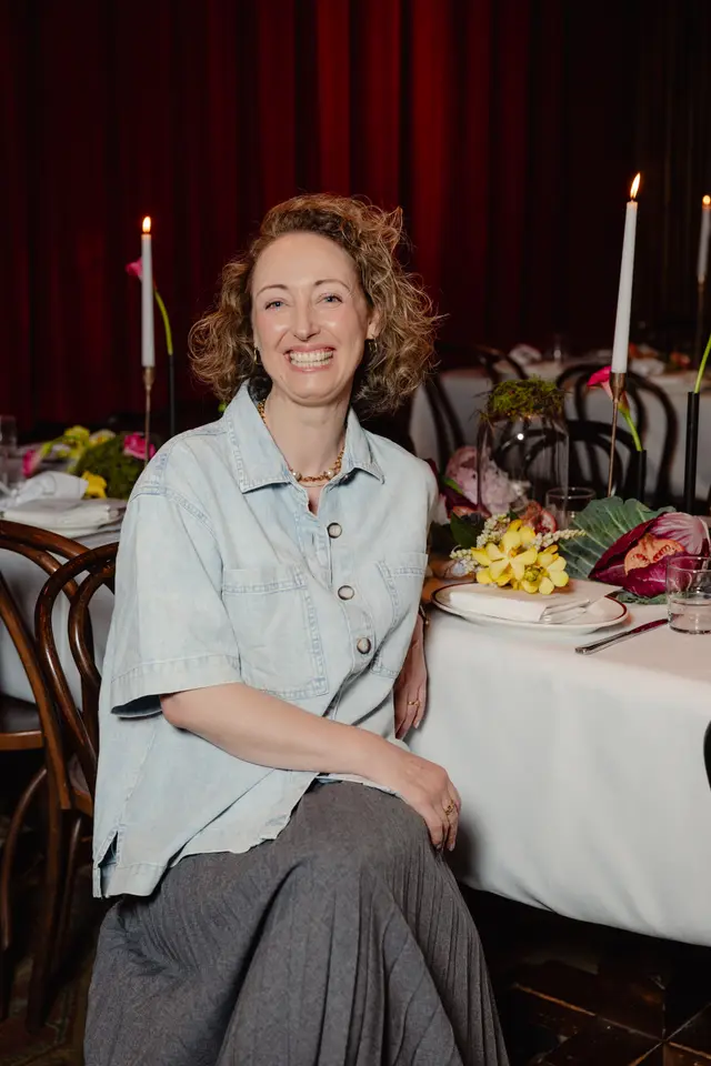 A smiling woman with curly hair sits at a beautifully set dinner table, looking directly at the camera. The table is adorned with elaborate floral arrangements, candles, and place settings, suggesting an elegant event. Red curtains form a warm backdrop, creating a sophisticated atmosphere.