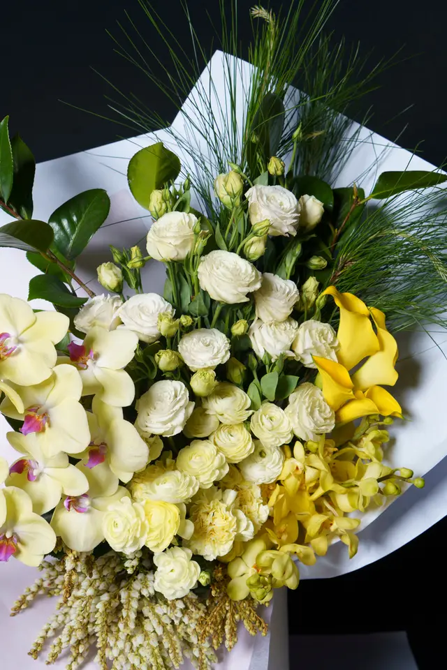 A vibrant, close-up studio shot of a large floral bouquet, presented against a dark background. The arrangement features a rich mix of cream, white, and various yellow flowers, including orchids, ranunculus, and calla lilies, interspersed with green foliage. The bouquet is wrapped in white paper, highlighting the fresh and diverse blooms.