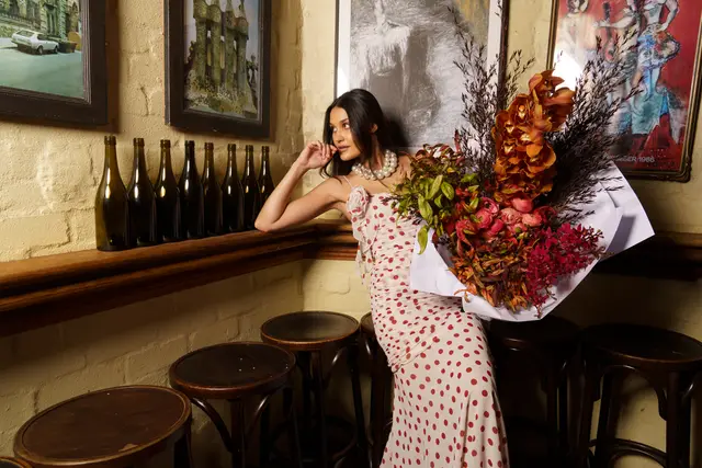 A woman in a white and red polka dot dress, adorned with a pearl necklace, leans against a wooden shelf while holding a large, vibrant bouquet of flowers. The bouquet features prominent orange orchids, red ranunculus-like blooms, and various green and dark foliage, wrapped in white paper. The setting is an indoor space with a light brick wall, dark wooden shelf lined with bottles, and framed artwork, creating a warm and styled atmosphere.