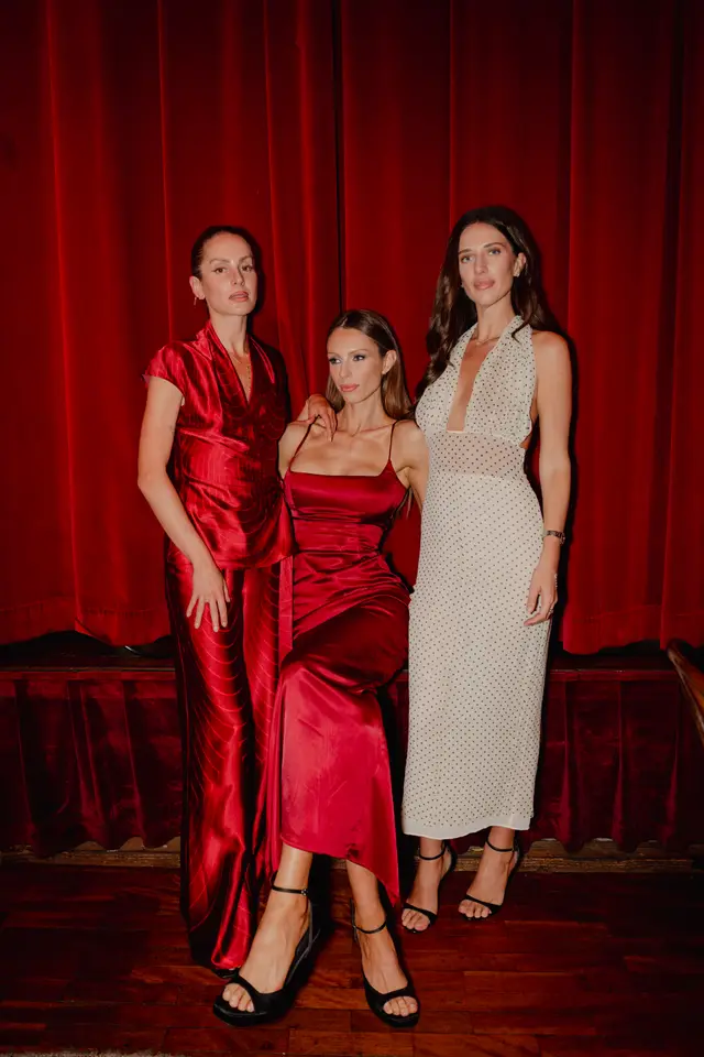 Three women are posing elegantly in formal attire against a rich, deep red velvet curtain backdrop. The woman on the left wears a red satin wrap dress, the central woman is seated in a red satin slip dress, and the woman on the right wears a cream polka dot halter-neck dress. They are all looking towards the camera, showcasing their outfits and accessories in a sophisticated setting.