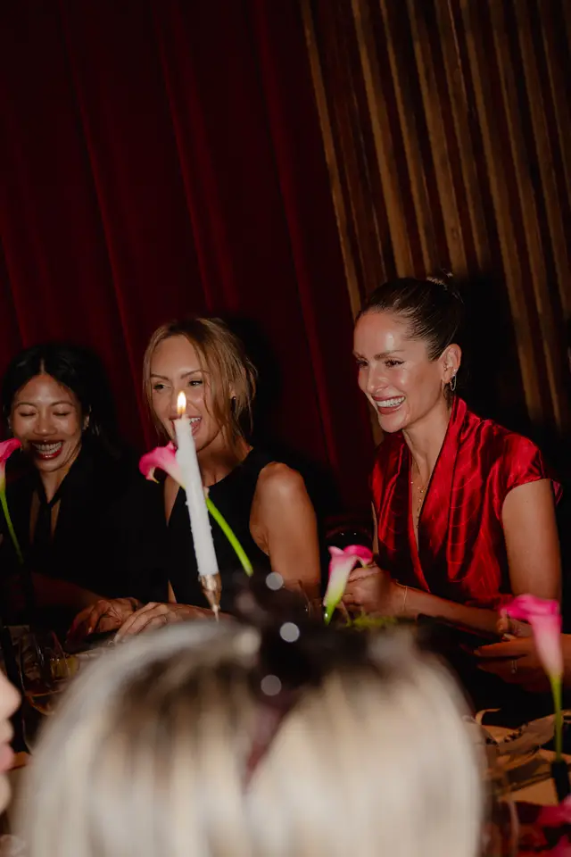 Three women are seated at a table, laughing and smiling, illuminated by warm light from a candle. Pink calla lilies are visible on the table, suggesting a celebratory or intimate event. The background features dark red curtains and wooden paneling.
