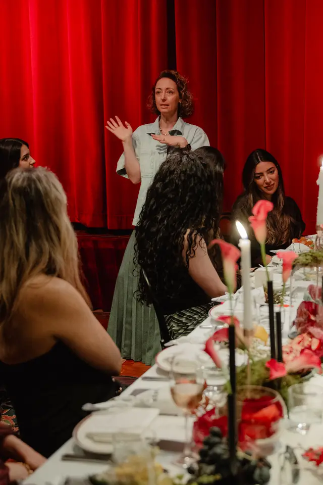 A woman with curly hair, wearing a light denim shirt and a green pleated skirt, stands and speaks with expressive hand gestures against a vibrant red curtain backdrop. She addresses a group of women seated at a long, elegantly set table adorned with candles, pink calla lilies, and other floral arrangements. The scene suggests an intimate event or dinner gathering with a speaker.