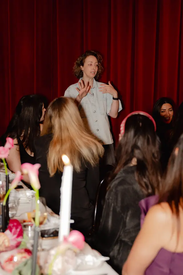 A woman with curly hair, wearing a light denim shirt, stands and speaks to a group of people seated at a table. She is gesturing with her hands, and her audience appears attentive. The background features rich, dark red curtains, and the foreground shows parts of a white tablecloth adorned with pink flowers and a lit candle.