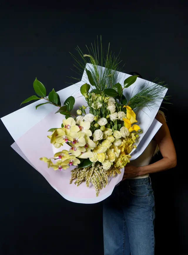 A person holds a large, vibrant floral bouquet featuring prominent yellow orchids, white spray roses, and yellow calla lilies, wrapped in white and light pink paper. The bouquet is presented against a dark, plain background, with the person's arm and lower body in blue jeans partially visible. The composition highlights the rich textures and colors of the flowers.
