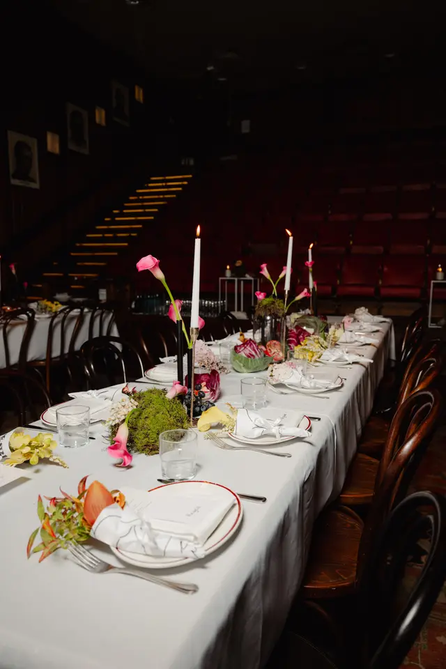A long dining table is elegantly set for an event, adorned with white tablecloths, place settings, and various floral and natural decorations. Lit candles provide a warm glow, highlighting pink and yellow calla lilies, green moss, and dark grapes. The background reveals a dimly lit venue with dark wooden chairs, red tiered seating, and framed pictures on a dark wall.