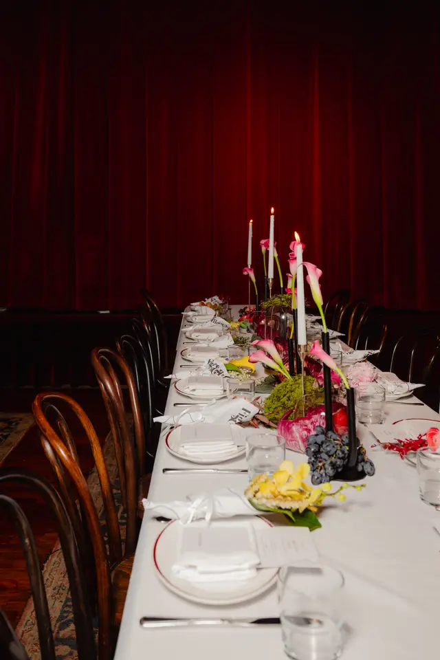 A long dining table is elegantly set for an event, featuring multiple place settings with white plates, folded napkins, and menus. The table is adorned with elaborate floral arrangements, including pink calla lilies, yellow orchids, and green moss, alongside lit candles in candelabras and bunches of dark grapes. The background consists of a dark red curtain, suggesting an indoor event space.