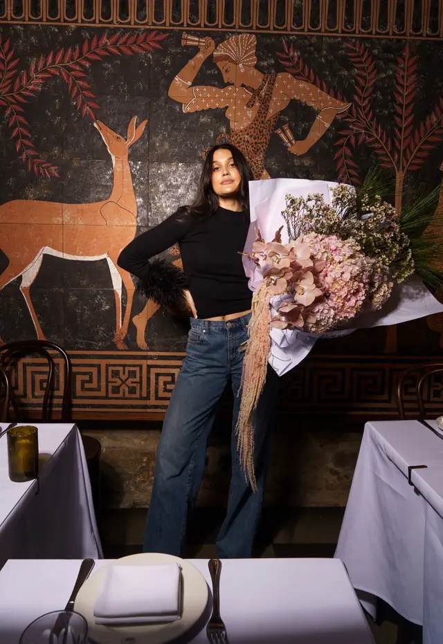 A full-length shot of a woman holding a large, elaborate bouquet of flowers, standing in a restaurant setting. She is wearing a black ribbed top with feathered cuffs and blue jeans. Behind her is a dark wall mural depicting ancient figures and animals in earthy tones.