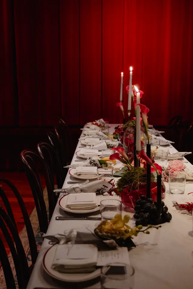 A long dining table is elegantly set for an event, featuring multiple place settings, lit candles, and elaborate floral arrangements. The scene is bathed in warm light, creating an intimate and luxurious atmosphere against a backdrop of rich red curtains. Various flowers, grapes, and decorative elements adorn the white tablecloth.