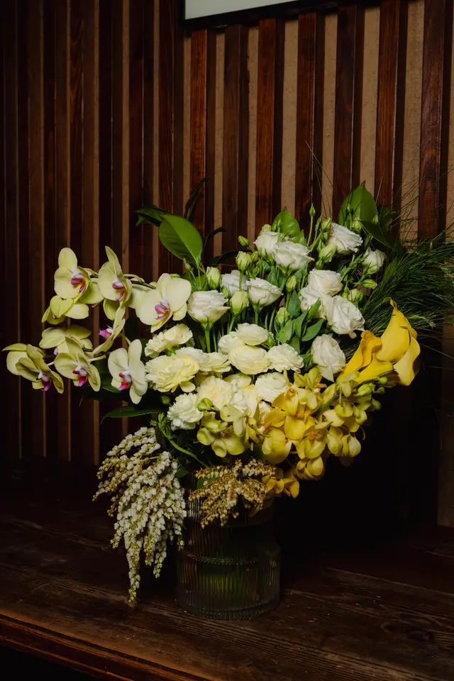 A large, elegant floral arrangement featuring yellow orchids, white and yellow ruffled flowers, and yellow calla lilies, displayed in a ribbed glass vase. The arrangement is set on a dark wooden surface against a dark slatted wooden wall, creating a sophisticated and rich visual.
