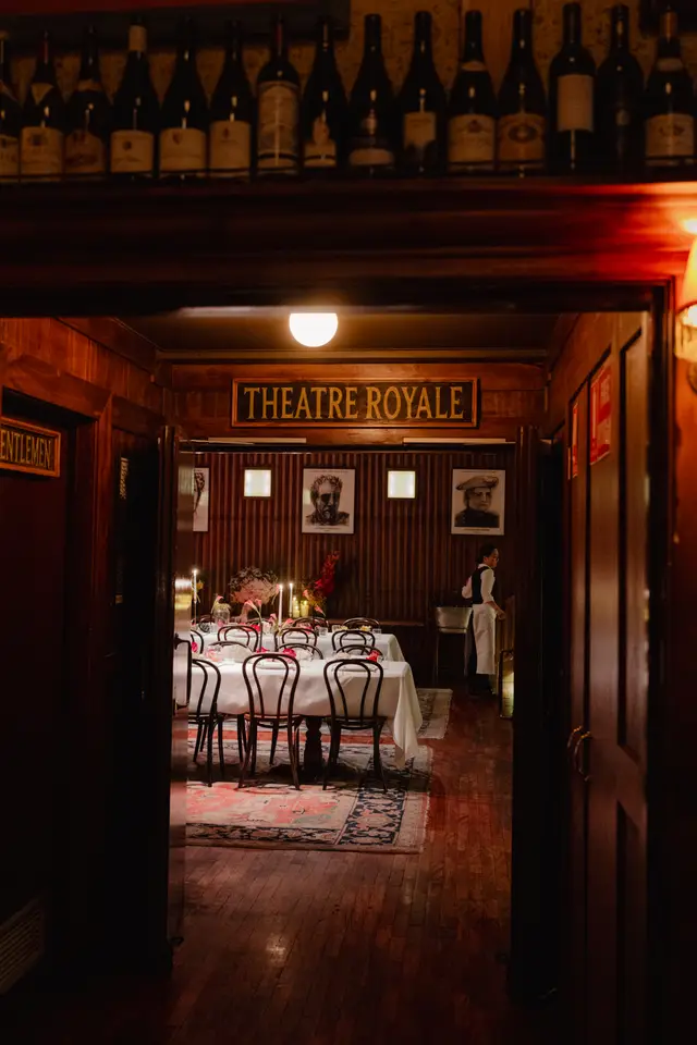 The image captures a view through a dark wooden doorway into a warmly lit dining room. A long table, set with white tablecloths, multiple lit candles, and floral arrangements featuring pink calla lilies and mixed pink/red flowers, is surrounded by dark bentwood chairs. In the background, a person in waitstaff attire stands near a silver bucket, and a prominent 'THEATRE ROYALE' sign is visible above the dining area entrance.