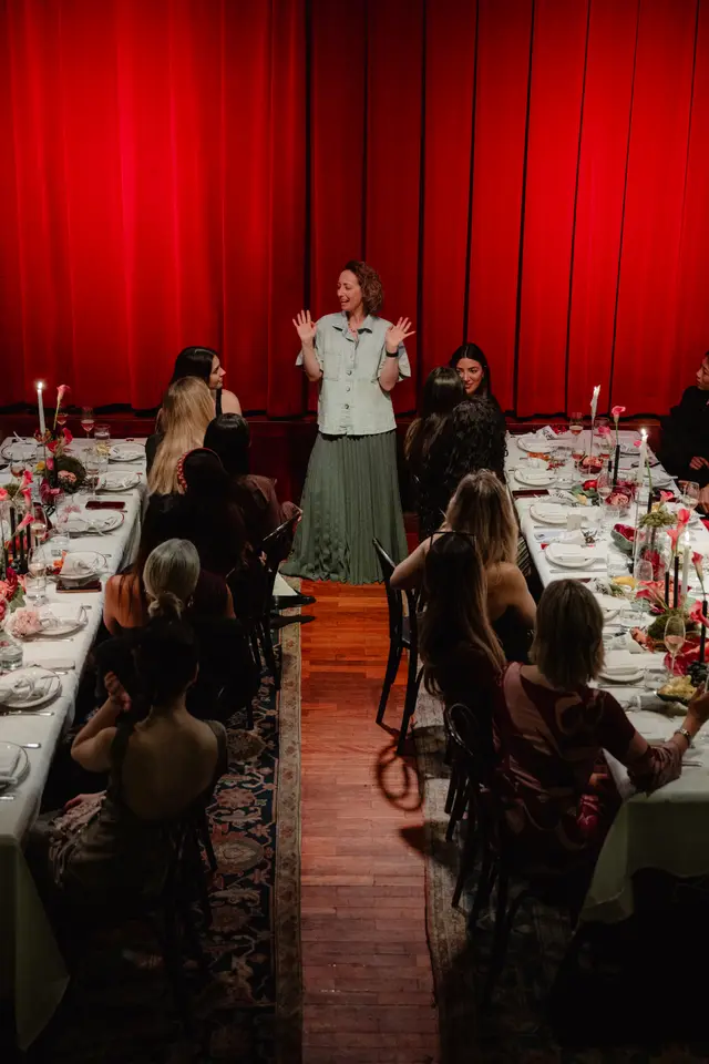 An overhead shot captures a woman speaking to a seated audience at a formal event. Two long dining tables, adorned with white tablecloths, candles, and floral arrangements, flank a central aisle where the speaker stands. A vibrant red curtain serves as the backdrop, creating a dramatic and intimate atmosphere for the gathering.