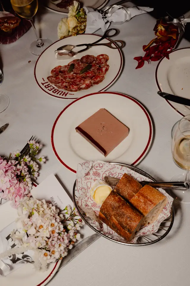 An overhead shot of an elegantly set dining table at an event, showcasing various appetizers including sliced charcuterie, a block of pâté, and a basket of fresh bread with butter. The table is adorned with multiple floral arrangements in shades of pink, white, yellow, and red, alongside a wine glass and a menu card.