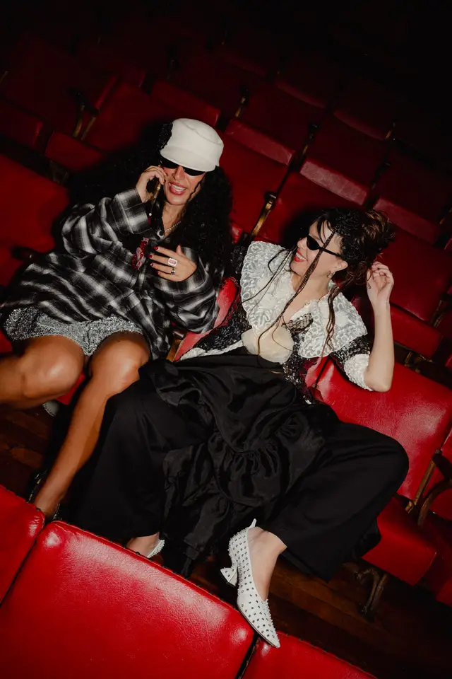Two women are seated in plush red theater seats, appearing joyful and engaged. One woman, wearing a plaid shirt and white cap, is on a phone call, while the other, in a sequined top and black wide-leg pants, smiles and touches her hair. The scene captures a candid moment within an event setting.