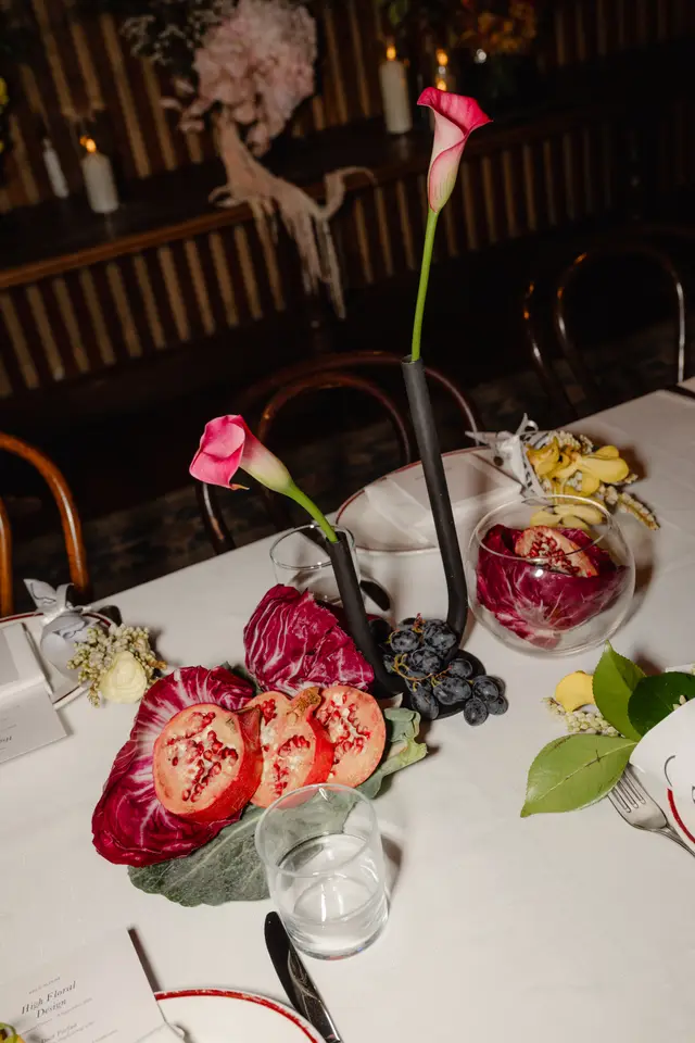 A close-up, overhead shot of an elegantly set dining table featuring a vibrant floral and fruit centerpiece. The arrangement includes pink calla lilies in black modern vases, halved pomegranates, radicchio, and dark grapes, alongside individual place settings with menus and smaller floral accents. The background shows blurred elements of a warm, dimly lit interior.