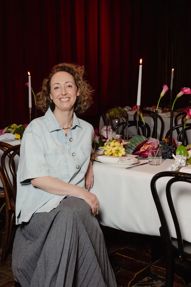 A smiling woman with curly hair is seated at a beautifully decorated event table. The table features elaborate floral arrangements, lit candles, and place settings, set against a backdrop of dark red curtains. The image captures a warm and inviting atmosphere.