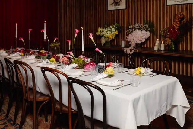 A long dining table is elegantly set for an event, featuring a white tablecloth, multiple place settings, and an elaborate centerpiece. The centerpiece includes various fresh flower arrangements, fruits like grapes, pomegranates, and lemons, and tall white candles in gold and black holders. The background shows dark wooden paneling, a dark red curtain, and a console table with additional large floral arrangements and candles.