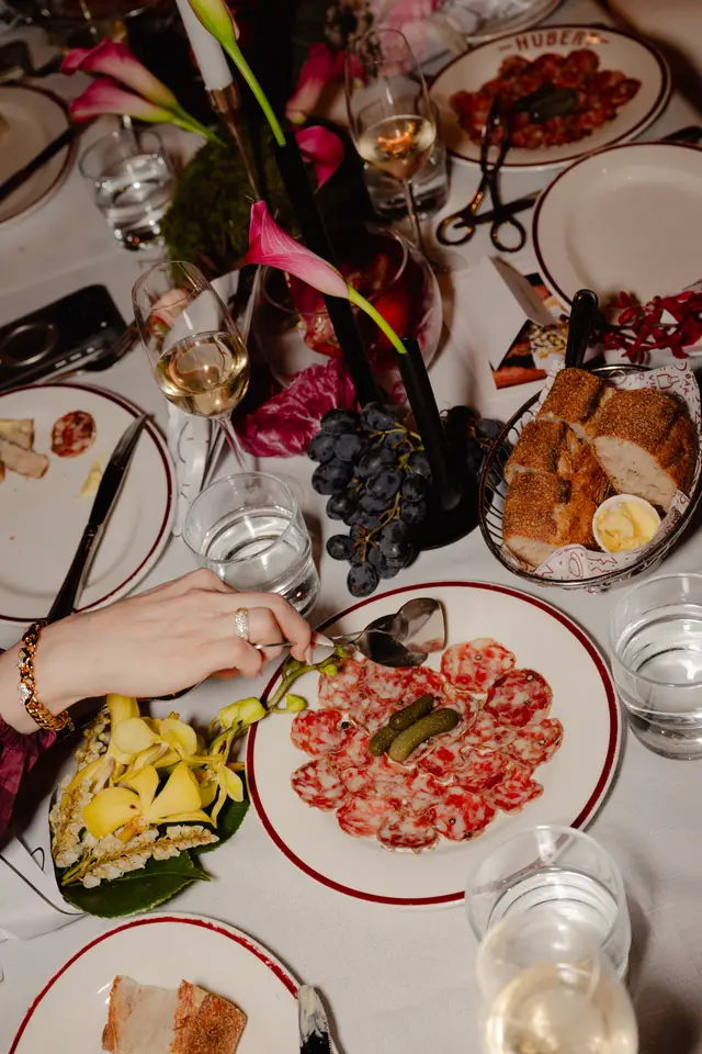 A high-angle shot captures an elegantly set dining table adorned with various dishes, drinks, and floral arrangements. A person's hand, wearing a gold bracelet and a diamond ring, is seen using serving tongs to pick from a plate of sliced cured meats. The table features white plates with red rims, wine and water glasses, a bread basket, grapes, and vibrant pink calla lilies and yellow orchids.