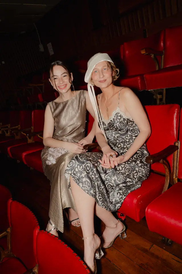 Two women are seated side-by-side in a row of red theater seats, looking towards the camera and smiling. The woman on the left wears a shimmering gold dress, while the woman on the right wears a patterned slip dress with lace trim and a white cloche hat. The background shows more empty red seats and a dark, dimly lit space.