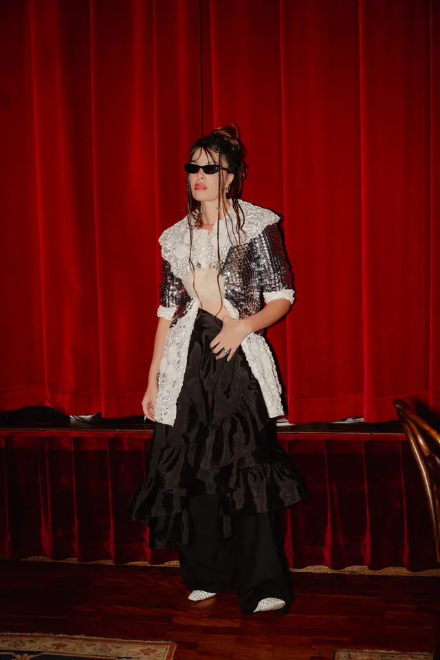 A full-body shot of a person standing in front of a deep red velvet curtain. The person wears a sequined jacket with a large white lace collar, black wide-leg pants, and white studded shoes, complemented by sunglasses and braided hair. The setting appears to be a stage or event space with a wooden floor and a partial rug.