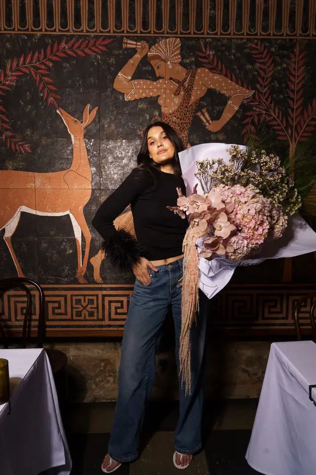 A full-length shot of a woman holding a large, elaborate bouquet of flowers, standing in front of a dark wall with an ancient-style mural. She is dressed in a black ribbed top with feathered cuffs, blue jeans, and white sandals. The setting appears to be an indoor space, possibly a restaurant or event venue, with tables draped in white cloths visible on either side.