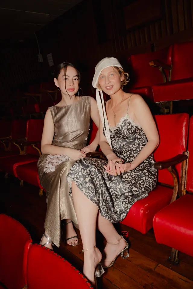 Two women are seated in an auditorium with rows of red seats and a dark wooden floor. The woman on the left wears a shimmering gold sleeveless dress, while the woman on the right wears an ornate patterned slip dress and a white cloche hat. They are captured in a candid moment, looking towards the camera with soft smiles.
