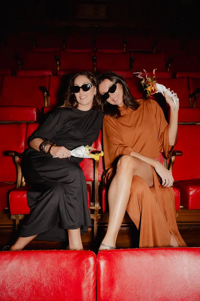 Two women wearing sunglasses and stylish dresses are seated in a row of red theatre seats, smiling at the camera. One woman holds a small bouquet of yellow and white flowers, while the other holds a bouquet with orange, red, and green flowers. The background shows more rows of empty red seats in a dimly lit setting.
