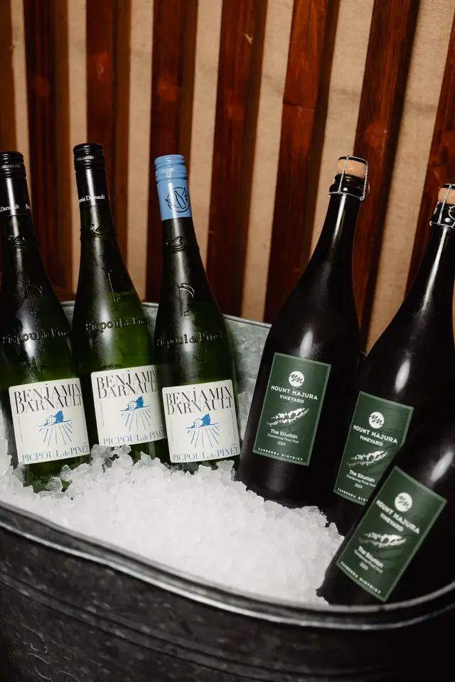 A close-up shot of several wine bottles chilling in a galvanized metal bucket filled with ice. Three green bottles with white labels and two dark bottles with green labels are visible, set against a rustic background of vertical wooden planks and burlap.