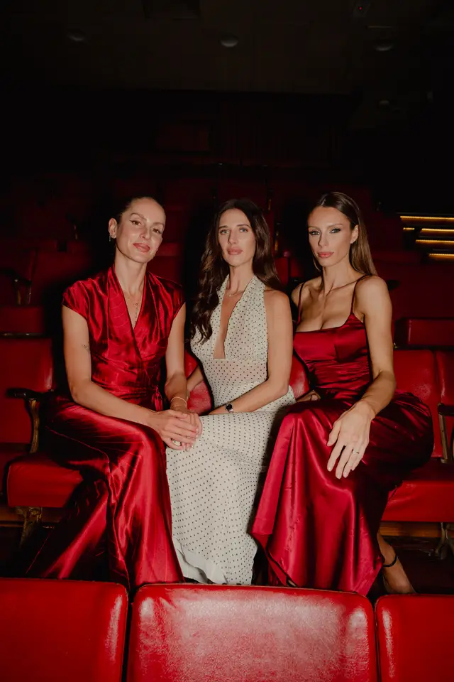 Three women are seated side-by-side in red theater seats, looking directly at the camera. Two women wear long red satin dresses, while the woman in the center wears a long white dress with black polka dots. The background is dark, suggesting a low-lit theater or event space.