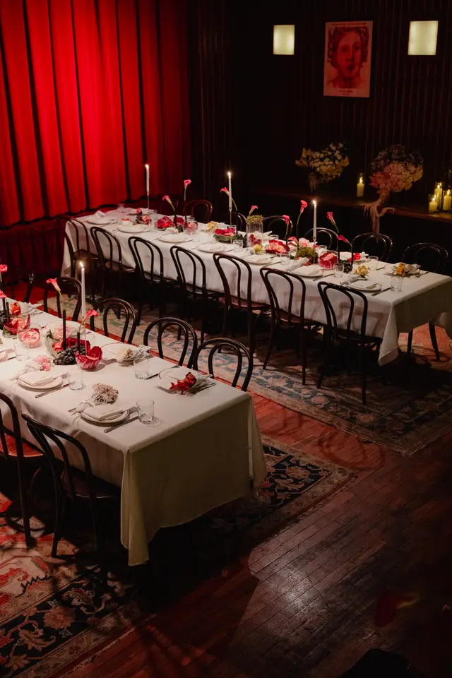 A high-angle shot captures two long dining tables elegantly set for an event in a dimly lit room. The tables are adorned with white tablecloths, candles, and elaborate floral and fruit centerpieces, creating a warm and inviting atmosphere. Red curtains line one wall, contrasting with dark wooden paneling and a patterned rug on the floor.