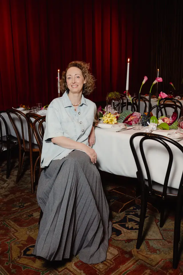 A smiling woman with curly hair is seated at a formally set event table, which is elaborately decorated with flowers, fruits, and lit candles. She is wearing a light blue button-up shirt and a long grey pleated skirt, with rich red velvet curtains and a patterned rug visible in the background.