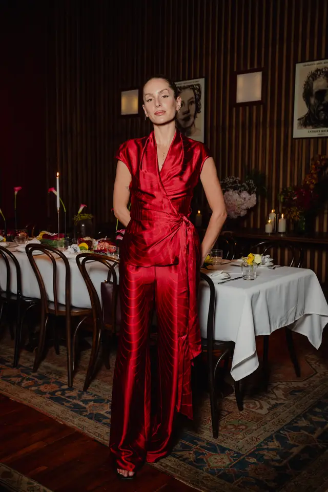 A full-body portrait of a woman wearing a striking red patterned wrap top and matching wide-leg pants. She stands in a dimly lit room with dark wood paneling, in front of a long dining table set with a white tablecloth, candles, and various floral arrangements. The setting suggests an elegant launch event or dinner.