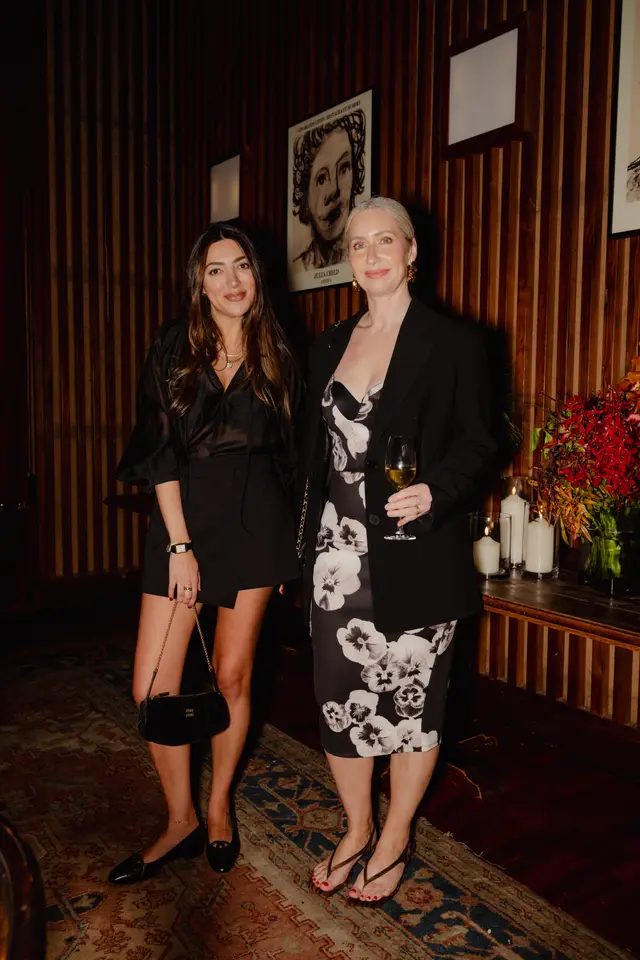 Two women are captured standing indoors at what appears to be an evening event. One woman wears a black dress and holds a Miu Miu bag, while the other wears a black and white floral dress and holds a wine glass. They are positioned in front of a dark wood-paneled wall adorned with framed artwork and a console table featuring lit candles and a vibrant floral arrangement.