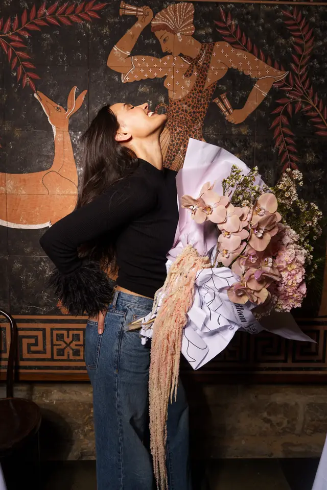 A woman with long dark hair, wearing a black ribbed top with furry cuffs and blue jeans, stands laughing with her head tilted back. She holds a large, elaborate bouquet featuring light pink orchids, pink hydrangeas, white filler flowers, and long, drooping light pink amaranthus, wrapped in pink and white paper. The background is a dark wall adorned with ancient Greek-style murals depicting a deer and a human figure in leopard print.