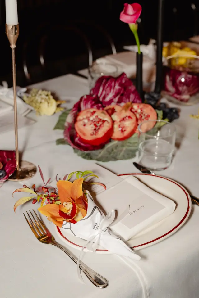 A close-up, high-angle shot of an elegantly set dining table, featuring a place setting with a white plate, a name card, a fork, and an orange orchid wrapped in decorative paper. The table is adorned with a white tablecloth, a gold candle holder, a water glass, and a centerpiece of halved pomegranates, radicchio, and dark grapes. A pink calla lily in a black vase is visible in the blurred background.