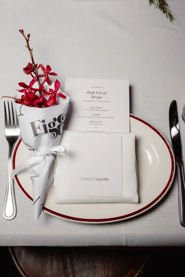 An elegant overhead shot of a single place setting at a formal event. The setting includes a white plate with a red rim, silver cutlery, a white linen napkin, a menu card, and a name card. A small bouquet of vibrant red orchids, wrapped in branded paper, is placed to the left of the plate.