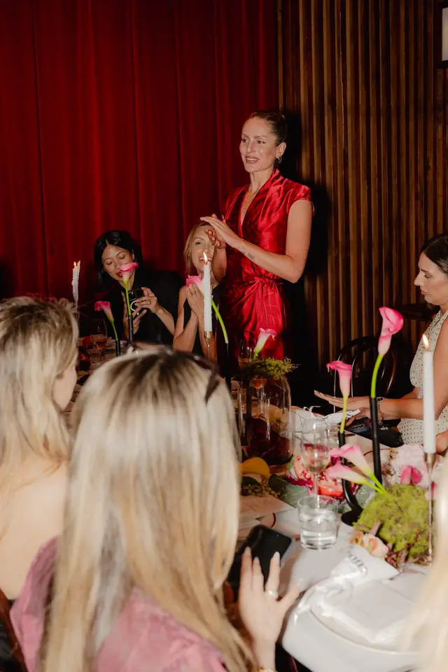 A woman in a vibrant red dress stands and speaks, gesturing with her hands, at a formal dining table. Several guests are seated around the table, which is elegantly decorated with pink calla lilies, lit candles, and other floral and fruit arrangements. The background features rich red curtains and dark wooden paneling, suggesting an upscale indoor event.