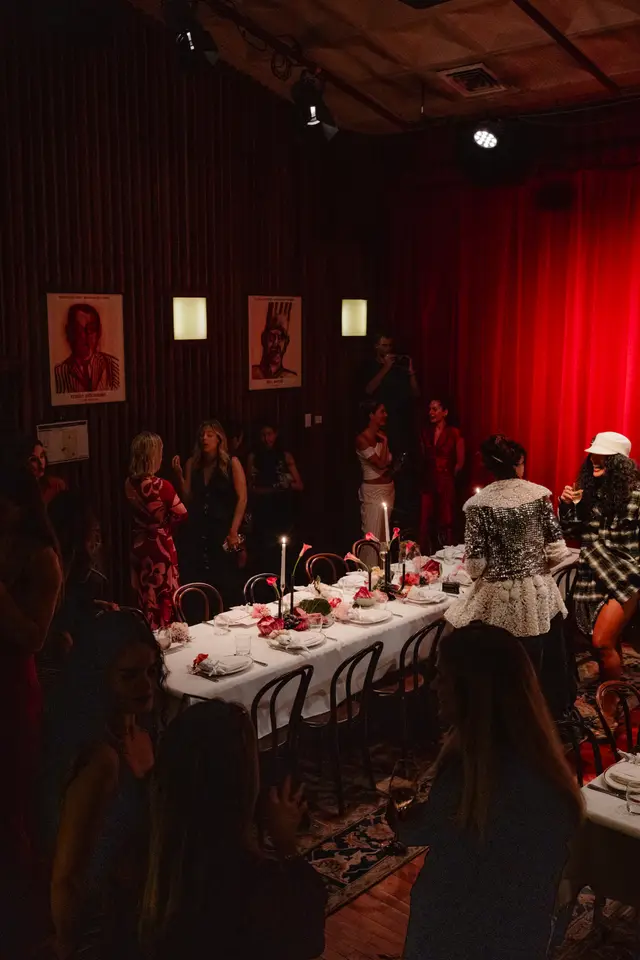 A dimly lit indoor event shows multiple people gathered around a long dining table set with a white tablecloth, candles, and floral arrangements. The room features dark wood paneling, wall art, and vibrant red curtains in the background, creating an intimate and festive atmosphere.