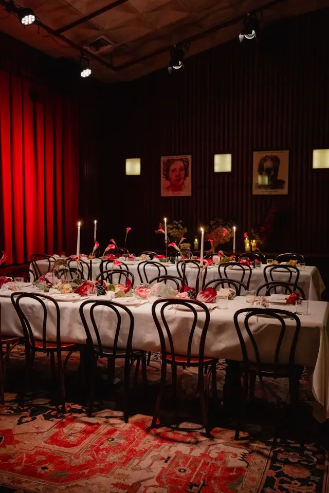 A long dining table is set for an event, adorned with white tablecloths, numerous floral arrangements, lit candles, and place settings. The room features dark wood-paneled walls, red curtains, and a patterned rug, creating an intimate and sophisticated atmosphere. Overhead spotlights illuminate the scene, highlighting the detailed table decor.